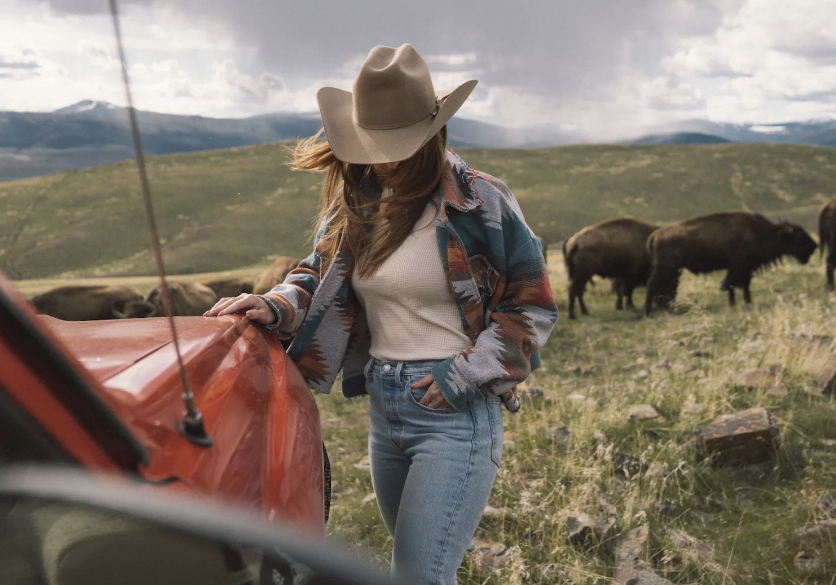 Woman walking towards a red car while wearing a light brown hat, muted colored jacket, and blue jeans with a bison heard in the background nearby mountains