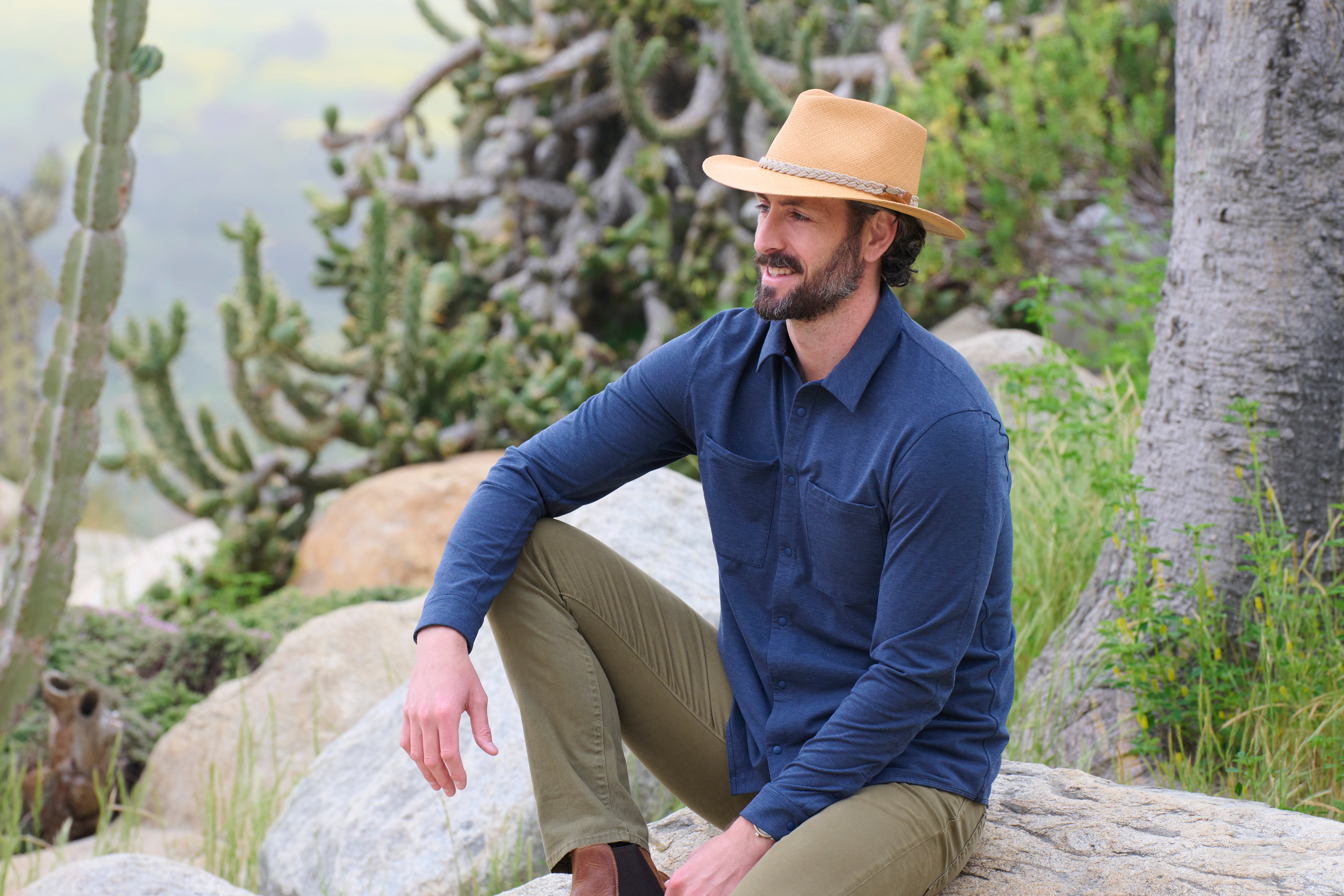 Bearded man wearing a sandy-colored hat with white band, blue long-sleeved buttoned up shirt, and light green pants smiling while sitting with his arm resting on his raised right knee on a rock out in the desert near many green cacti.
