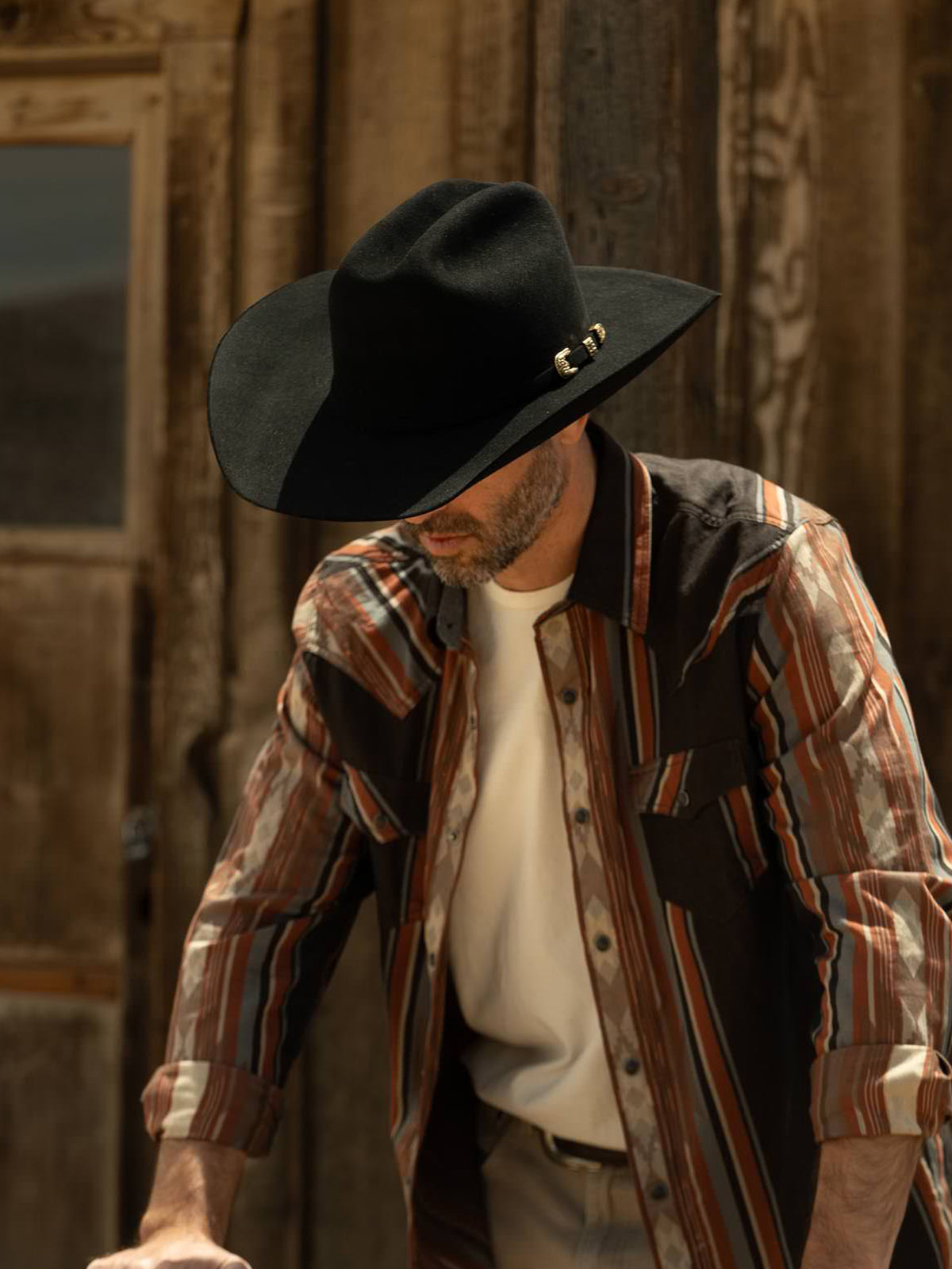 Man wearing a black cowboy hat and patterned shirt leaning against a wooden structure