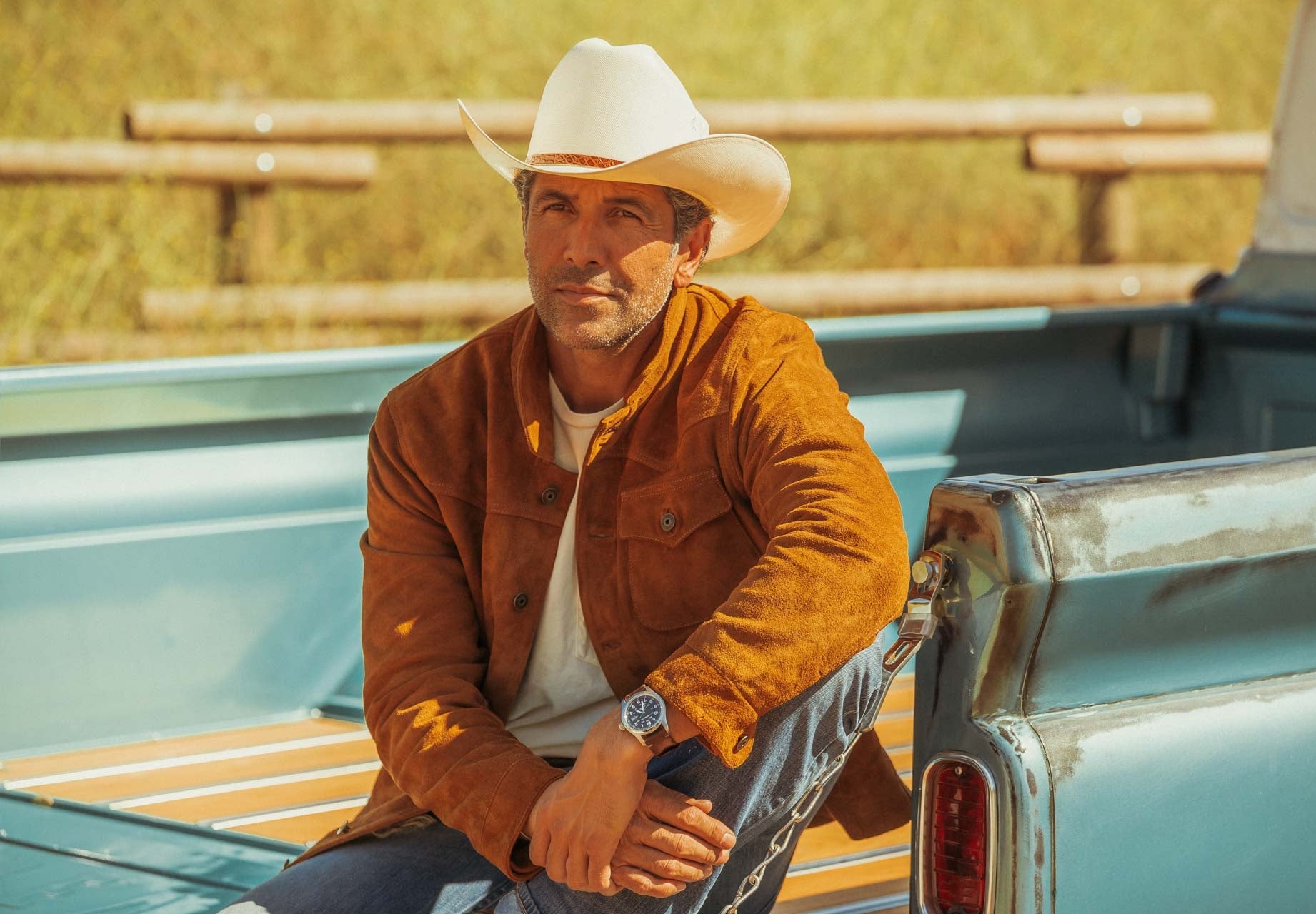 Man wearing a cowboy hat and brown jacket sitting on the back of a truck in a field.