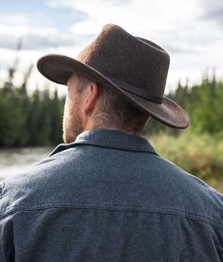 Man looking away wearing a brown long brim hat and dark blue jacket looking towards a river with trees along the way