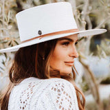 Woman wearing a white hat with light brown band and white sweater with small holes smiling against an old small tree