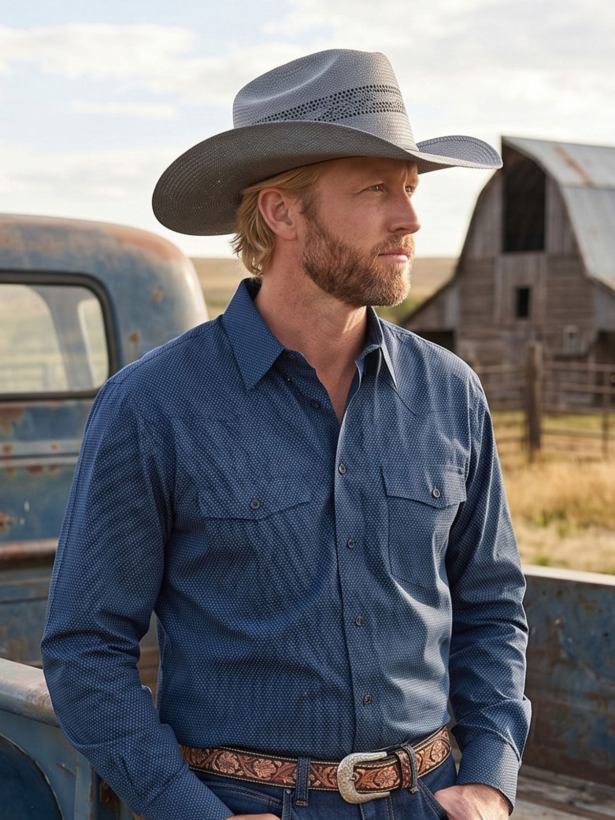 Bearded man wearing a gray straw cowboy hat sitting on the back of a rusty blue truck near a farmhouse out in the dry fields