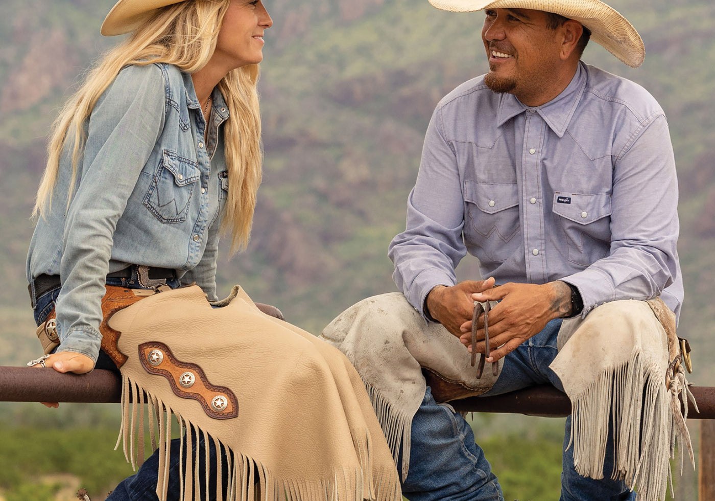 Two people in cowboy hats sitting on a fence with mountains in the background