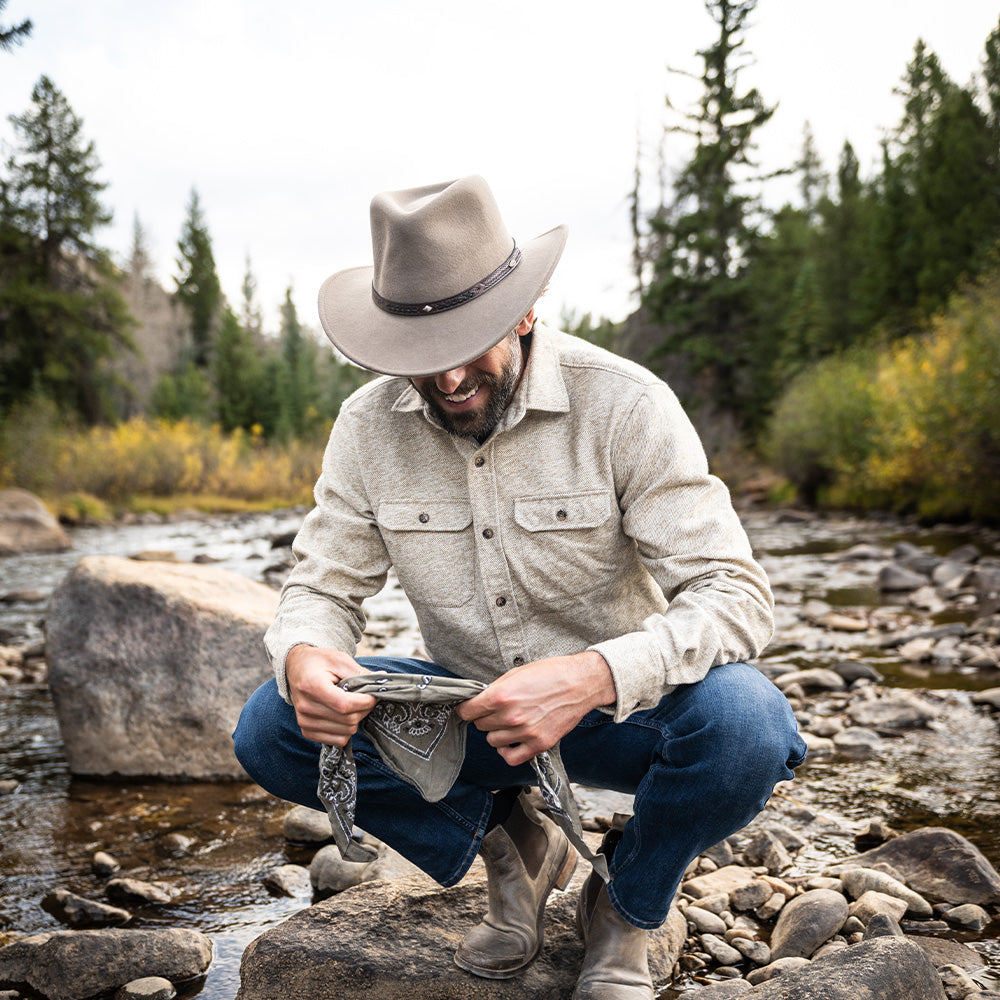 Smiling man standing on a rock in the middle of a forested rocky stream while holding a gray and black bandana wearing blue jeans, tan long sleeve button shirt, and tan hat with brown band