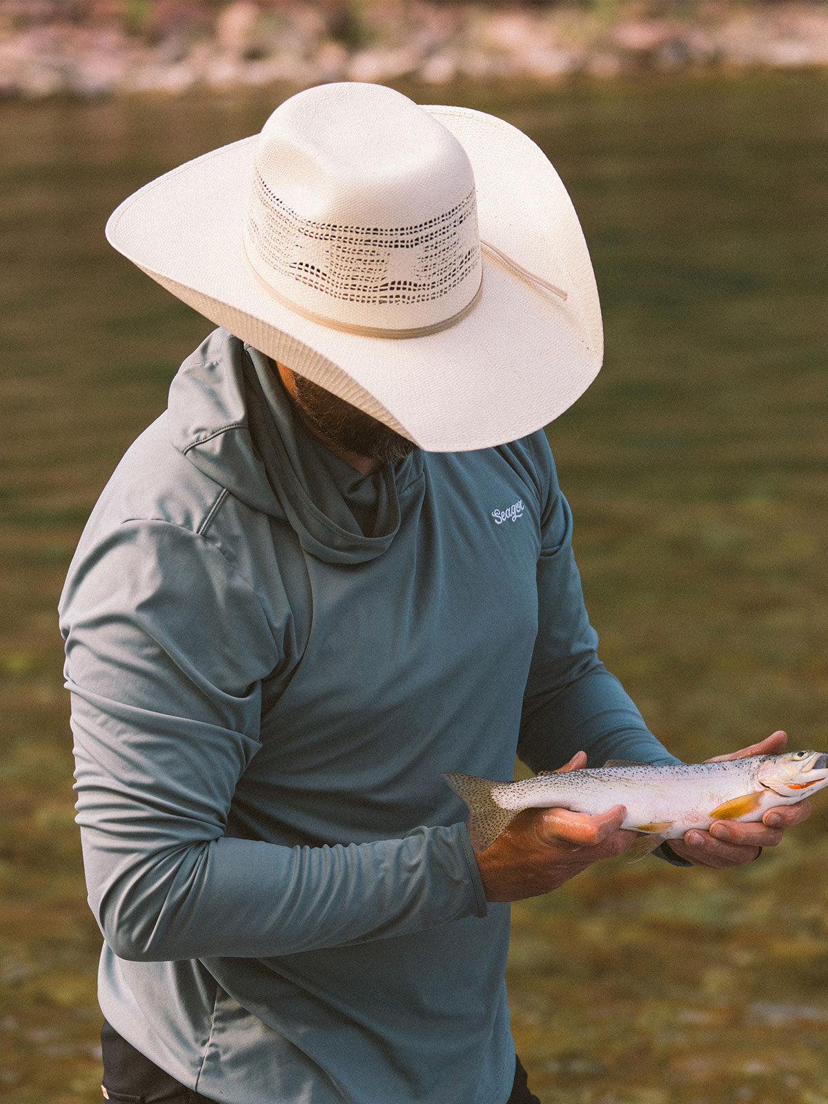 Man wearing a natural-colored cowboy hat and blue long-sleeved shirt while standing in a river with his head down