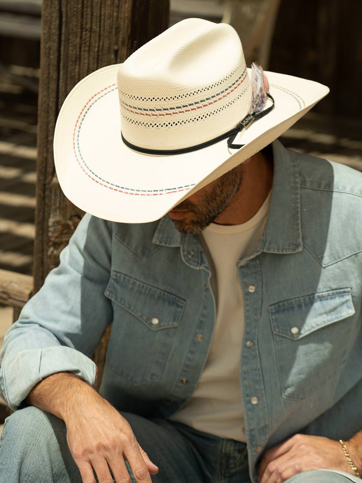 Man wearing a white cowboy hat and denim jacket sitting against a wooden post