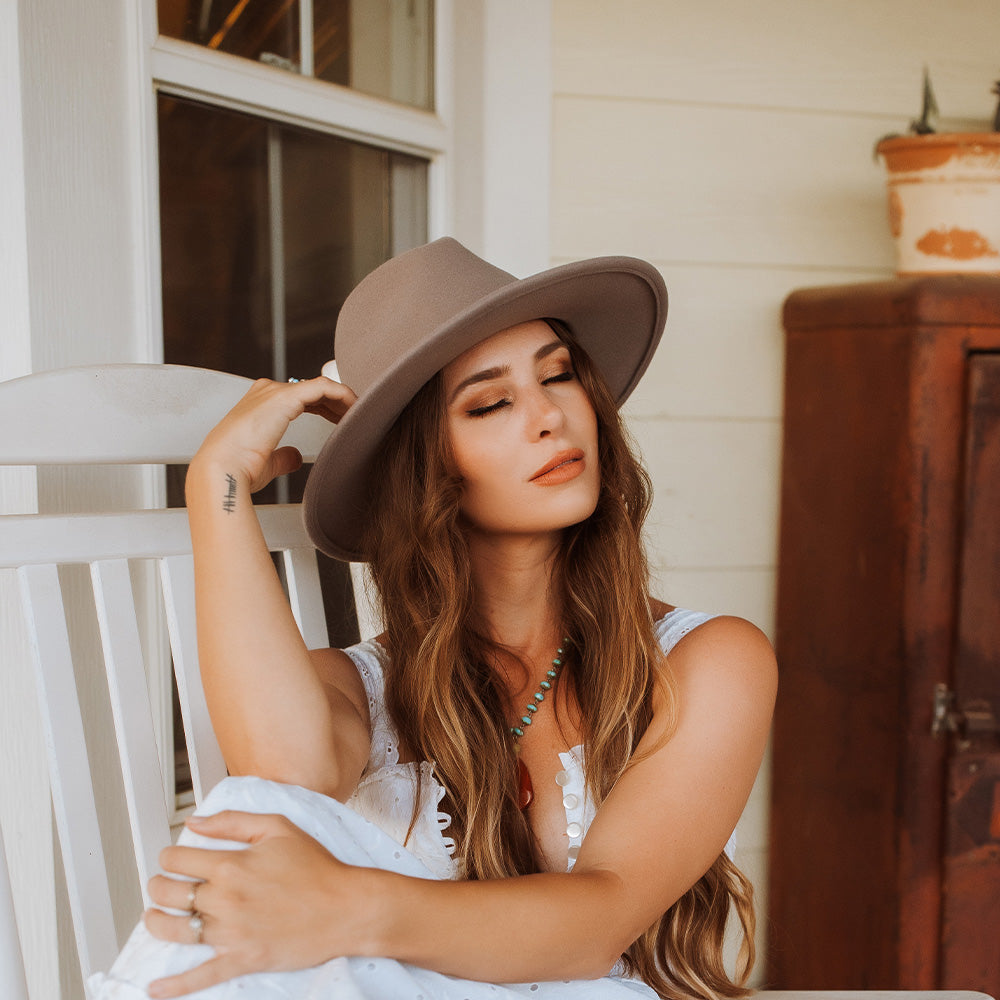 Woman resting her eyes leaning on a chair against a white house and window while wearing a light brown hat and white dress